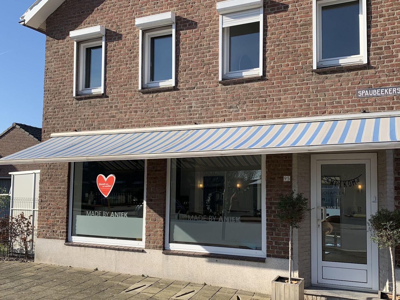 De gevel van Made by Aniek: een bakstenen huis met gestreepte luifel en twee ramen met de naam van de salon